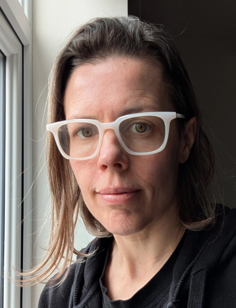 A close-up portrait of a person with long hair and glasses, looking directly at the camera, with natural light coming from a nearby window.